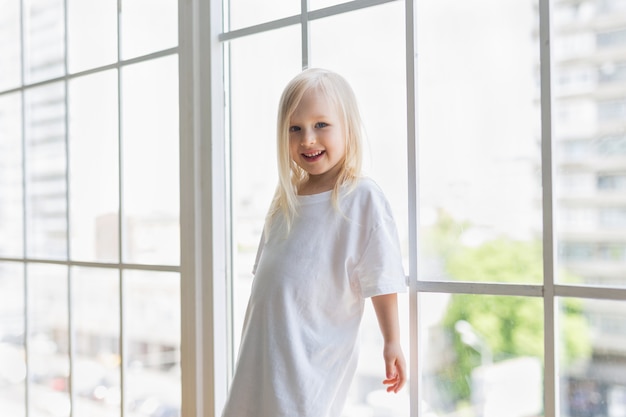 Happy pretty girl wearing blank white dress near big window