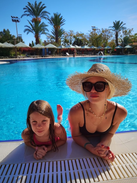 Happy pretty little girl with mother in big straw hat when they are resting at swimming pool of spa hotel.