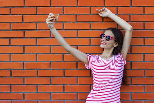 Happy pretty smiling woman makes self-portrait on smartphone over colorful red brick background