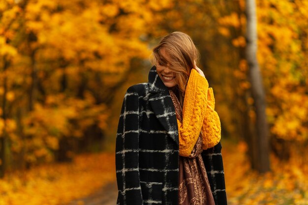 Happy pretty smiling woman with hair in fashion clothes with black coat and yellow knitted sweater enjoy autumn weather in amazing golden park