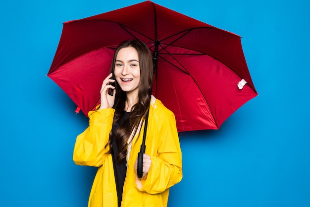 Happy pretty smiling young woman with umbrella using smartphone in autumn day over colorful blue wall