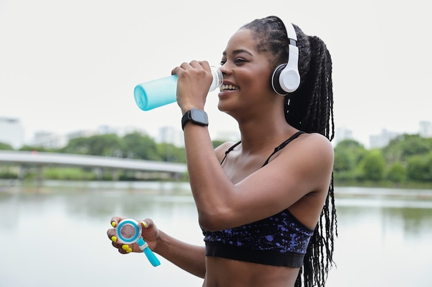 Happy pretty young Black fit woman in heaphones drinking fresh water after training outdoors