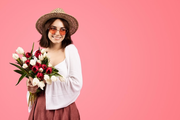 Happy pretty young caucasian woman in casual hat and glasses with flower bouquet enjoy birthday