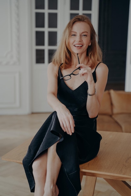 Happy pretty young female wearing alluring black dress holds glasses sitting on table at home