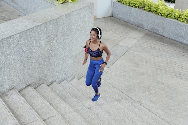 Happy pretty young Vietnamese sportswoman in headphones running up the stairs