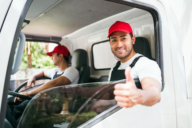 赤い帽子をかぶったアシスタントと幸せなプロのトラック運転手親指を立てて笑顔でカメラを見る