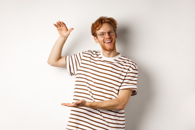 Happy and proud redhead guy with glasses showing something big, shaping large object with hands over white background, smiling at camera