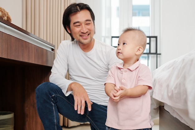 Happy proud young father looking at his adorable little son standing on the floor