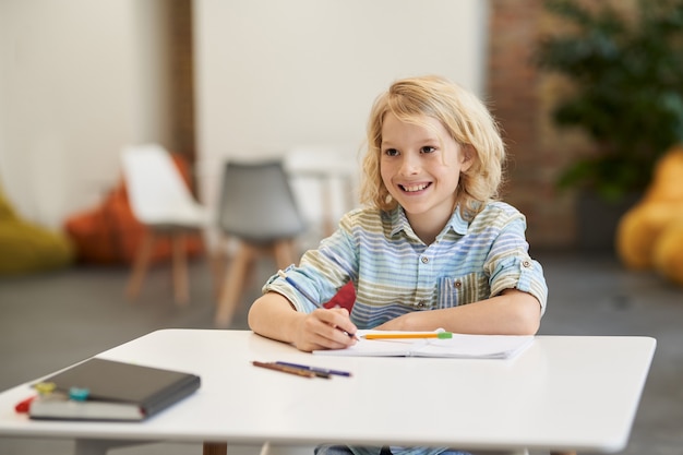 Happy pupil joyful school boy smiling away while writing in his notebook sitting at the desk in