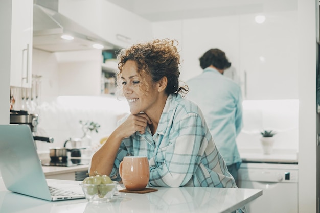 Happy real couple enjoy time together in the kitchen at home Woman using and watching laptop computer and man washing dishes working in background Relationship and living man and woman in apartment