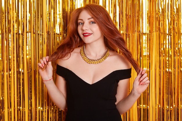 Happy red haired female with red lips looking at camera with flirting facial expression, touching her curls, wearing black dress and necklace