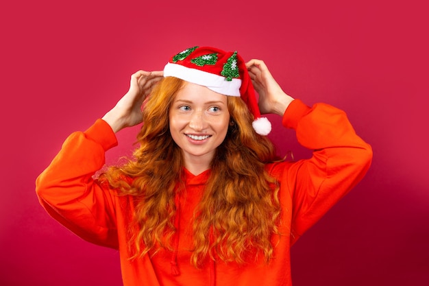 Happy red-haired girl, with a bright smile and gorgeous curly hair, in a Santa Claus hat on a red wall. Holiday concept. 