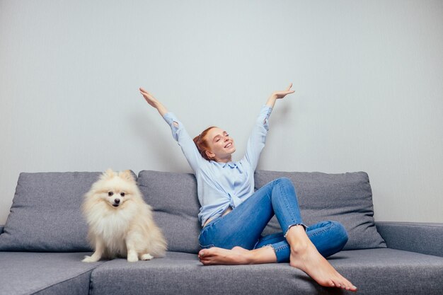 Happy, redhaired ginger woman in shirt and jeans satting with her white fluffy spitz have a rest on gray new folding sofa copyspase.