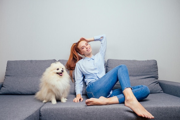 Happy redhaired ginger woman in shirt and jeans satting with her white fluffy spitz have a rest on gray new folding sofa copyspase