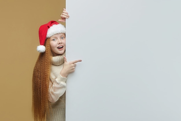 Happy redhaired girl in a Santa hat with a white Billboard