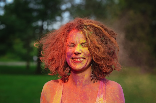 Foto felice donna dai capelli rossi che indossa una maglietta bianca in posa ricoperta di vernice colorata secca holi al parco