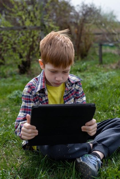 Happy redhead boy playing tablet or watching cartoons while sitting on green grass in the backyard in the village summer holidays closeup