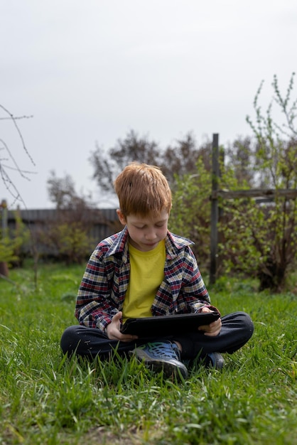 Happy redhead boy playing tablet or watching cartoons while sitting on green grass in the backyard in the village vacation