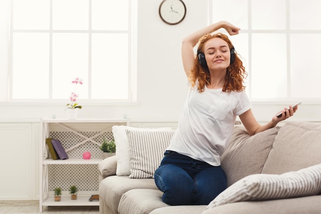 Happy redhead girl listening to music online on smartphone in headphones. Curly woman having rest at home, leisure and technology concept