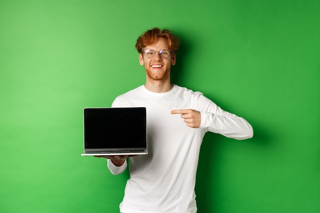 Фото Счастливый рыжий парень в очках и белой футболке с длинным рукавом, указывая пальцем на пустой экран ноутбука и улыбаясь, стоя на зеленом фоне.