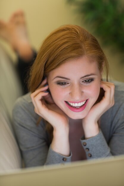 Happy redhead lying on the sofa using her laptop at night