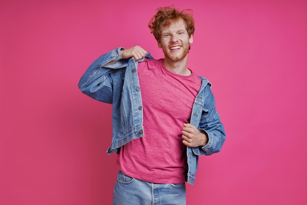 Felice uomo rosso in abbigliamento denim guardando la fotocamera mentre si trova su sfondo rosa