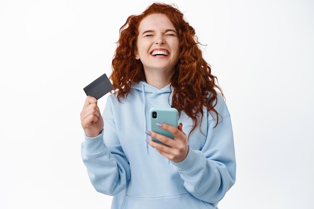 Happy redhead woman rejoicing, holding smartphone and plastic credit card, close eyes and laugh from overjoy, standing  on white, online shopping