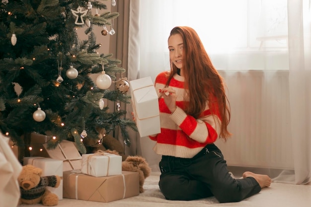 Happy redhead woman in sweater with gifts under Christmas tree with lights New Year holidays