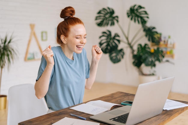 Happy redhead young businesswoman is working laptop and getting good message at remote work place at the desk in home office Exited girl goes crazy because of good news from the internet