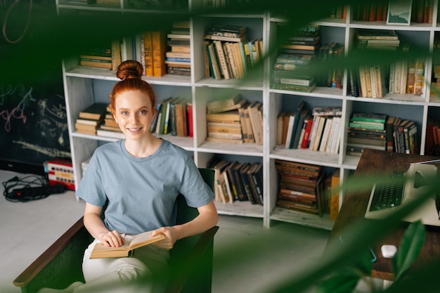 Photo happy redhead young woman student is reading book enjoys of rest at home office looking at camera cute lady enjoying books at library room on background of bookshelves