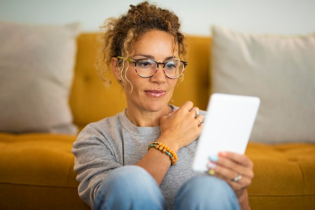 Happy relaxed adult cute woman use electronic device reader to read an ebook and enjoy her indoor leisure activity at home Pretty female people wearing glasses and smile Portrait of lady