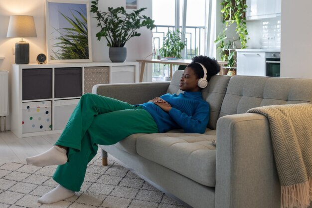 Photo happy relaxed african american woman in headphones closing eyes enjoying music sits on sofa