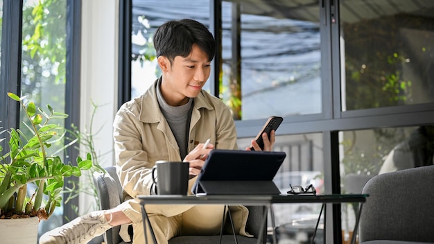 コーヒー ショップのリモート作業で彼の携帯電話とポータブル タブレットを使用して幸せでリラックスしたアジア人