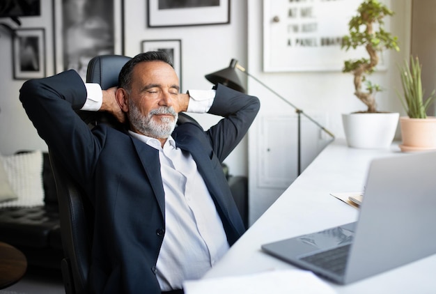 Happy relaxed european senior businessman in suit and closes eyes resting alone at table with