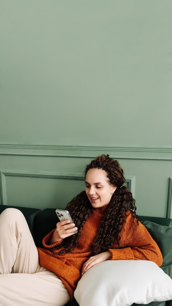 Счастливая расслабленная молодая женщина, сидящая на диване с помощью мобильного телефона, улыбающаяся леди, смеющаяся, держа смартфон