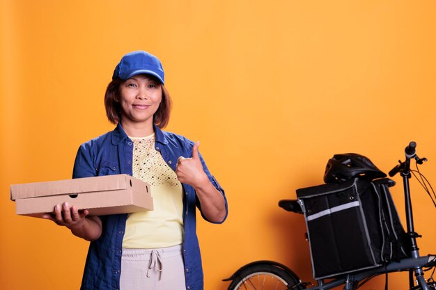 Happy restaurant courier in blue uniform doing aprproval sign\
holding takeout food boxes, delivering pizza order to client.\
pizzeria worker bringing orders with bike. take out food service\
and concept