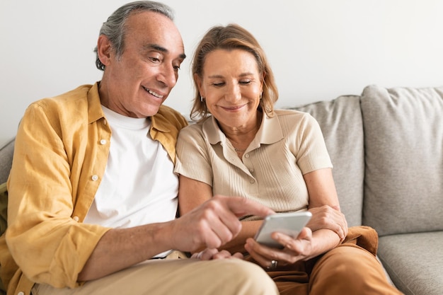 happy retired family spouse watching funny photos on cellphone sitting on sofa at home free space
