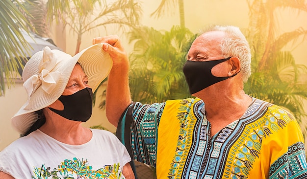 安全のためにマスクを着用して幸せな退職者。引退した老人が老婦人の帽子の下を覗き、妻を見ます。