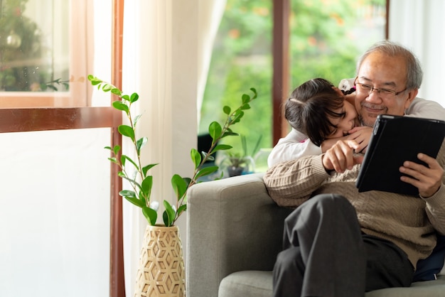 Happy retirement elderly man sitting on sofa at living room with granddaughter using digital tablet together.