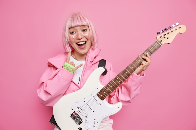 Happy rock star makes horn heavy metal sign being member of popular band or famous solo artist poses with acoustic electric guitar has trendy pink hair wears fashionable clothes poses indoor