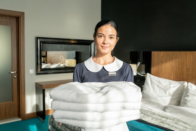 happy room service staff holding stack of white clean sheets in bedroom