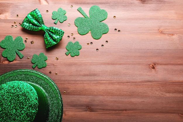 Happy Saint Patrick's Day greeting card with traditional symbols, shamrock, green attire. Green hat, bow tie, St Patricks Day shamrocks, golden confetti on wooden background, copy space, top view