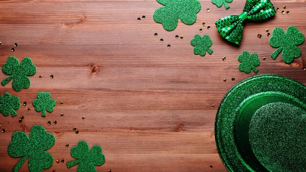 Happy Saint Patrick's Day greeting card with traditional symbols, shamrock, green attire. Green hat, bow tie, St Patricks Day shamrocks, golden confetti on wooden background, copy space, top view