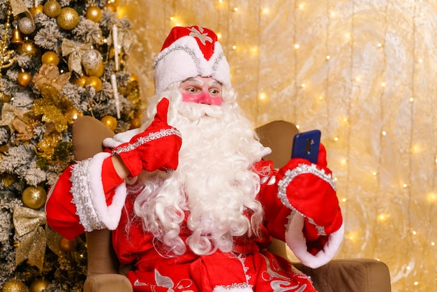 Счастливый Санта-Клаус держит смартфон, используя мобильное приложение для социального общения на расстоянии, наблюдая ...