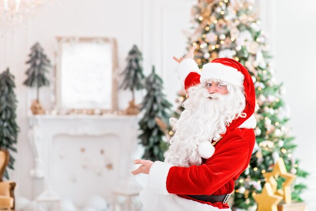 Happy Santa Claus indoors pointing to christmas tree