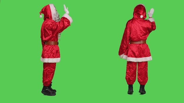 Happy santa claus wave hello in studio, wearing festive costume with glasses and red hat over full body greenscreen. Saint nick greeting someone and spreading holidays spirit.