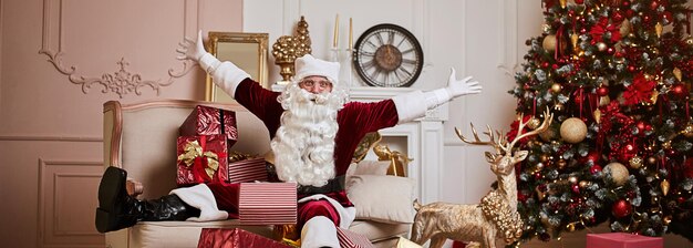 Happy santa claus with gifts near fireplace and christmas tree new year and merry christmas concept