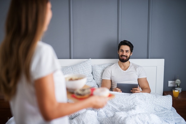 Happy satisfied bearded man looking at his girlfriend or wife brings breakfast in the bed while he reading news from a tablet at home or hotel.