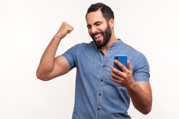 Happy satisfied man with beard in blue shirt holding smartphone and smiling making yes gesture celebrating online lottery or giveaway victory Indoor studio shot isolated on white background