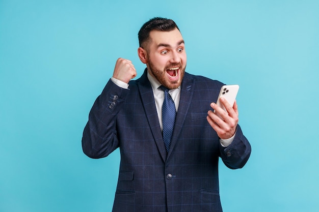 Счастливый довольный мужчина с бородой в официальном костюме держит смартфон и улыбается, делая жест «да», празднуя онлайн-лотерею или победу в розыгрыше. Внутренний студийный снимок на синем фоне